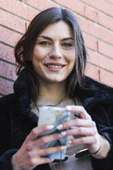 Smiling woman holding mobile phone against brick wall - PNAF00655