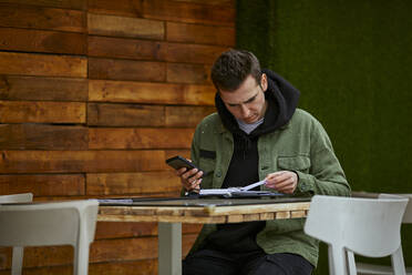 Man with mobile phone checking list while sitting at table by wall - ACPF01121