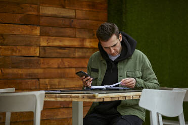 Mann mit Mobiltelefon, der eine Liste überprüft, während er an einem Tisch an der Wand sitzt - ACPF01121