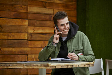 Smiling man writing in diary while talking on mobile phone at table in sidewalk cafe - ACPF01122