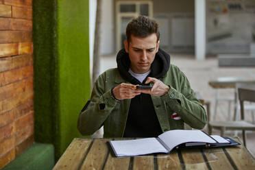 Handsome man taking photo of diary while sitting at table in sidewalk cafe - ACPF01126