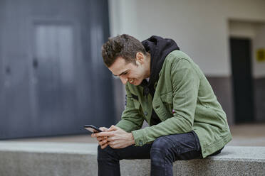 Smiling handsome man using smart phone while sitting on retaining wall at sidewalk cafe - ACPF01128