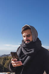 Junger Mann in warmer Kleidung, der ein Mobiltelefon gegen den blauen Himmel im Winter benutzt - MGRF00162