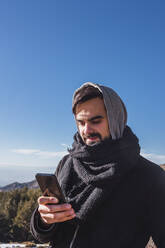 Young man in warm clothing using mobile phone against blue sky during winter - MGRF00162