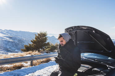 Mann benutzt Mobiltelefon, während er sich im Winter auf die Motorhaube eines Fahrzeugs stützt - MGRF00166