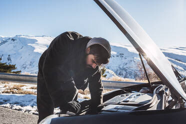Man repairing car on snow covered land against sky during winter - MGRF00172