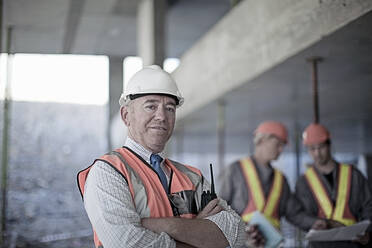 Selbstbewusster männlicher Bauunternehmer mit verschränkten Armen auf einer Baustelle mit Bauarbeitern im Hintergrund - AJOF01032