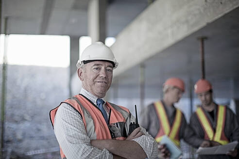 Confident male contractor with arms crossed at site with construction workers seen in background - AJOF01032