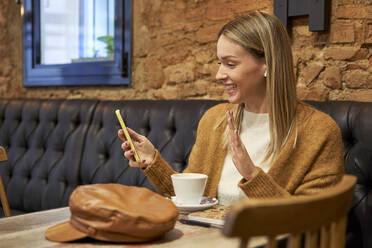 Lächelnde Frau führt einen Videoanruf über ihr Smartphone durch, während sie in der Cafeteria sitzt - VEGF03879