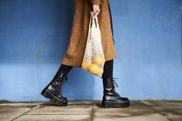 Frau, die Orangen in einer wiederverwendbaren Tasche trägt, während sie auf dem Fußweg an einer Mauer vorbeigeht - VEGF03898