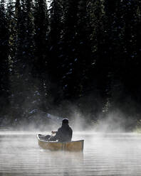 Mann paddelt Kanu auf Teich im Nebel, umgeben von dunklem Wald - CAVF93138