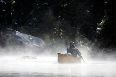 Bärtiger Mann mittleren Alters paddelt Boot auf einem nebligen See in Kanada - CAVF93140