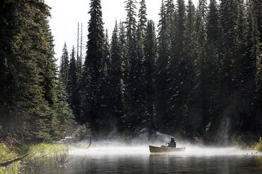 Man paddling boat on a remote lake on a foggy misty day surrounded by trees - CAVF93143