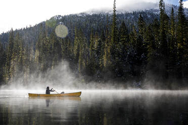 Man paddles canoe on lake by mountains with mist and fog on sunny day - CAVF93145