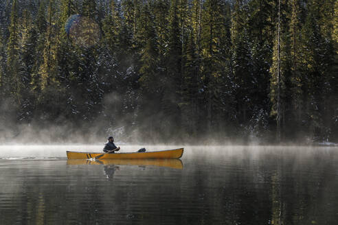 Mann in einem Kanu auf einem nebligen See an einem sonnigen Tag, umgeben von Bäumen - CAVF93146