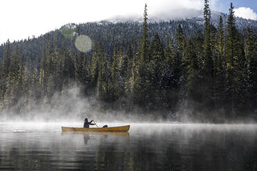 Man paddles canoe on lake with mist and fog on sunny day by forest - CAVF93147
