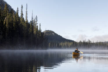 Young man paddles canoe on misty calm lake with sky - CAVF93149