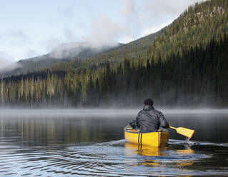 Rückansicht eines Mannes, der mit einem Boot auf einem ruhigen, nebligen See in Kanada paddelt - CAVF93151