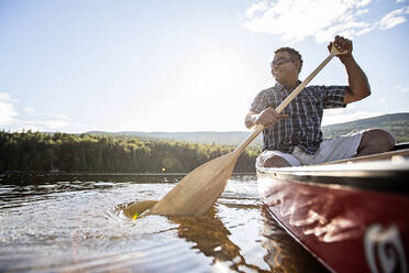 Lächelnder afroamerikanischer Mann in rotem Kanu taucht das Paddel in einen See in Maine - CAVF93160