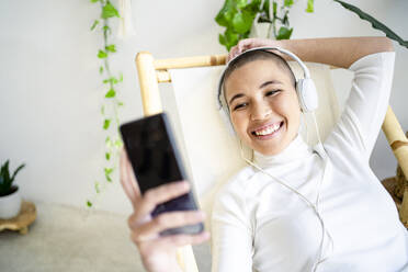 Junge Frau benutzt ihr Smartphone, während sie zu Hause über Kopfhörer Musik hört - RCPF00696