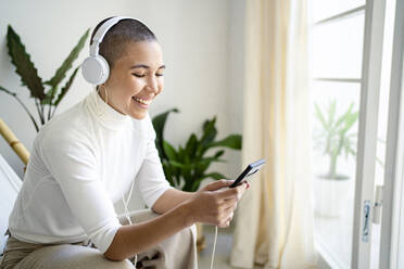 Woman using mobile phone while listening music through headphones in living room - RCPF00697