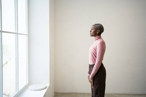 Woman looking through window while standing by wall at home - RCPF00723