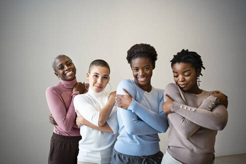 Smiling female friends self hugging against wall at home - RCPF00728