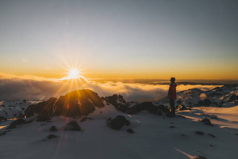 Junge Frau mit roter Jacke beobachtet den Sonnenuntergang am Horizont im Whakapapa Ski Resort, Neuseeland - CAVF93254