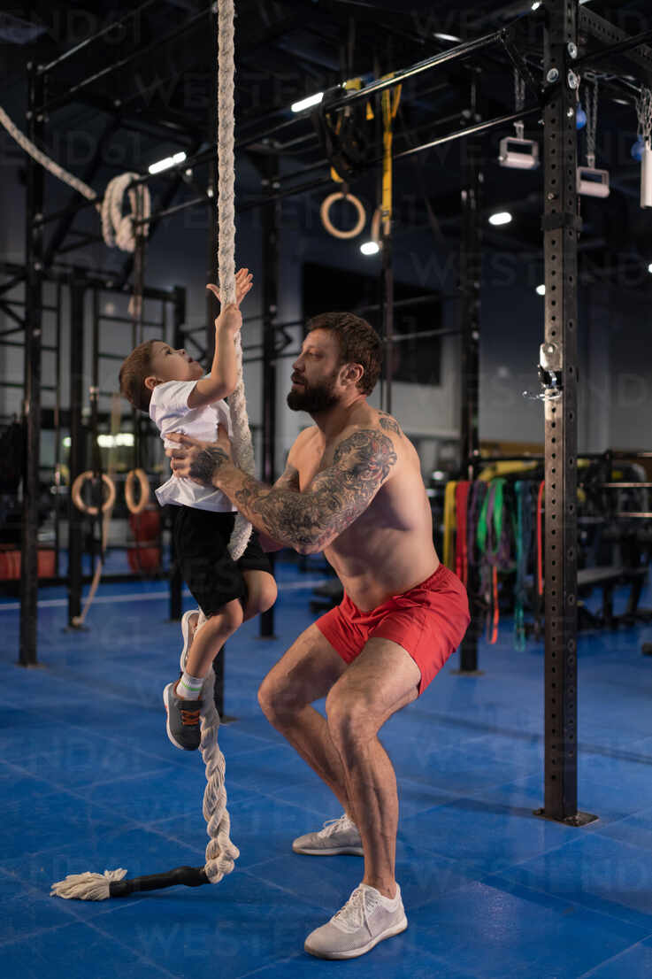 Shirtless father teaching boy to climb rope during training stock