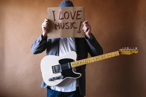 Mature male guitarist holding paper with text against wall - JCMF01893
