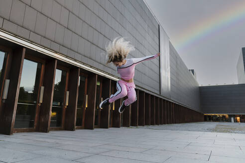 Cheerful woman jumping on footpath against building - MTBF00851