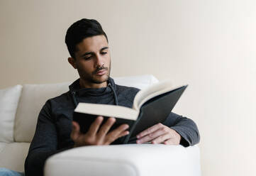 Young man reading book while sitting on sofa at home - EGAF01731