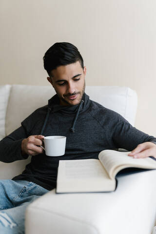 Junger Mann mit Kaffeetasse, der zu Hause auf dem Sofa sitzt und ein Buch liest, lizenzfreies Stockfoto