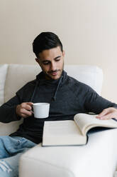 Young man with coffee cup reading book while sitting on sofa at home - EGAF01732