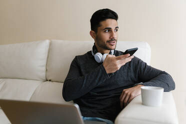 Young man talking through smart phone while sitting on sofa at home - EGAF01735