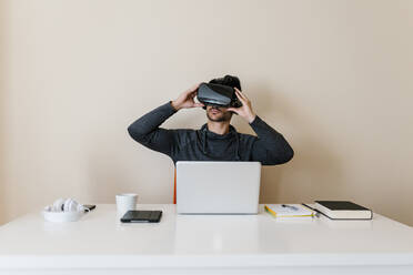 Junger Mann trägt einen Virtual-Reality-Simulator, während er am Schreibtisch im Wohnzimmer sitzt - EGAF01744