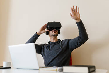 Man wearing virtual reality simulator with arm raised while sitting at table in living room - EGAF01745