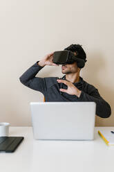 Man sitting with laptop wearing virtual reality simulator at table in living room - EGAF01746