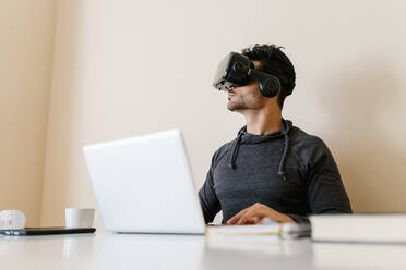 Young man sitting with laptop wearing virtual reality simulator in living room - EGAF01747