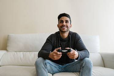 Lächelnder junger Mann sitzt auf dem Sofa und spielt zu Hause ein Videospiel - EGAF01748