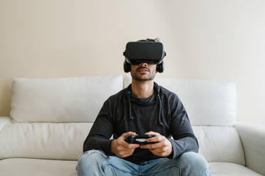 Young man wearing virtual reality simulator playing video game while sitting on sofa at home - EGAF01750