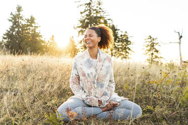 Fröhliche Frau sitzt mit gekreuzten Beinen im Gras und macht Yoga bei Sonnenuntergang - AKLF00035