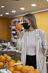 Frau mit Gesichtsschutz, die in einem Geschäft während der Pandemie orangefarbene Früchte untersucht - VEGF03918