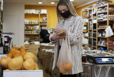 Frau mit Schutzmaske schüttet während der COVID-19 in einem Geschäft Orange in eine Tasche - VEGF03919