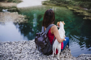 Woman with backpack embracing dog while sitting on rock at lake - EBBF02468