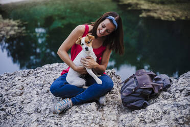 Playful woman holding dog while sitting on rock at lake - EBBF02469