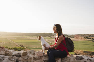 Smiling woman holding dog while sitting on rock against sky - EBBF02470