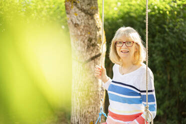 Happy woman sitting on swing in garden - AKLF00038