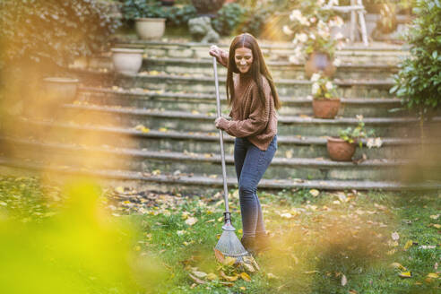 Smiling woman sweeping with rake in garden - AKLF00045