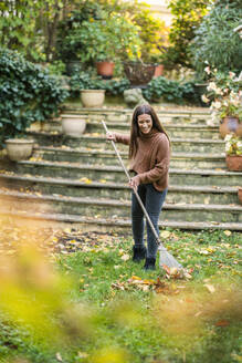 Woman in warm clothing sweeping with rake in back yard garden - AKLF00046